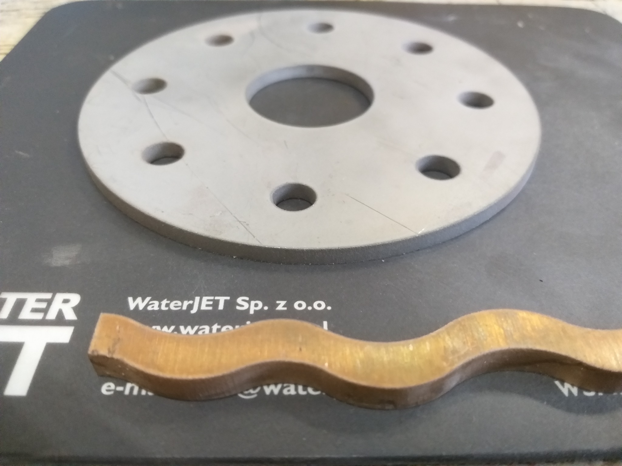 Obrabiany metalowy element w kształcie falistej listwy z mosiądzu oraz okrągły element z otworami na czarnym podłożu z nadrukiem WaterJET.
