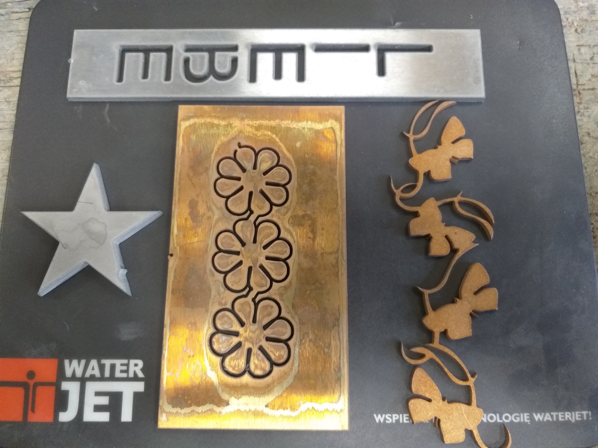 Wycięte elementy z metalu: gwiazda, fragment z literami, płytka z wyciętymi kwiatami oraz dekoracyjny motyw roślinny, wykonane technologią waterjet.