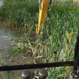 Widok z kabiny żółtej koparki JCB podczas czyszczenia rowu z roślinności, w tle pole ze słomą w balotach.