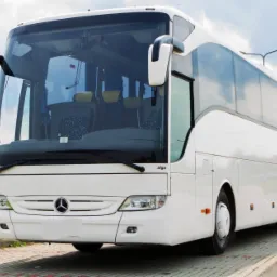 Biały autokar Mercedes Tourismo z widocznymi fotelami pasażerów przez przednią szybę, stojący na kostce brukowej w słoneczny dzień.