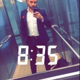 Mężczyzna w garniturze robi selfie w nowoczesnej windzie z lustrzanymi ścianami i cyfrowym zegarem 8:35 na ekranie telefonu.