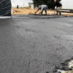 Wyrównywanie świeżo wylanego betonu na placu budowy, dwóch pracowników w roboczych ubraniach, widoczny samochód dostawczy w tle.