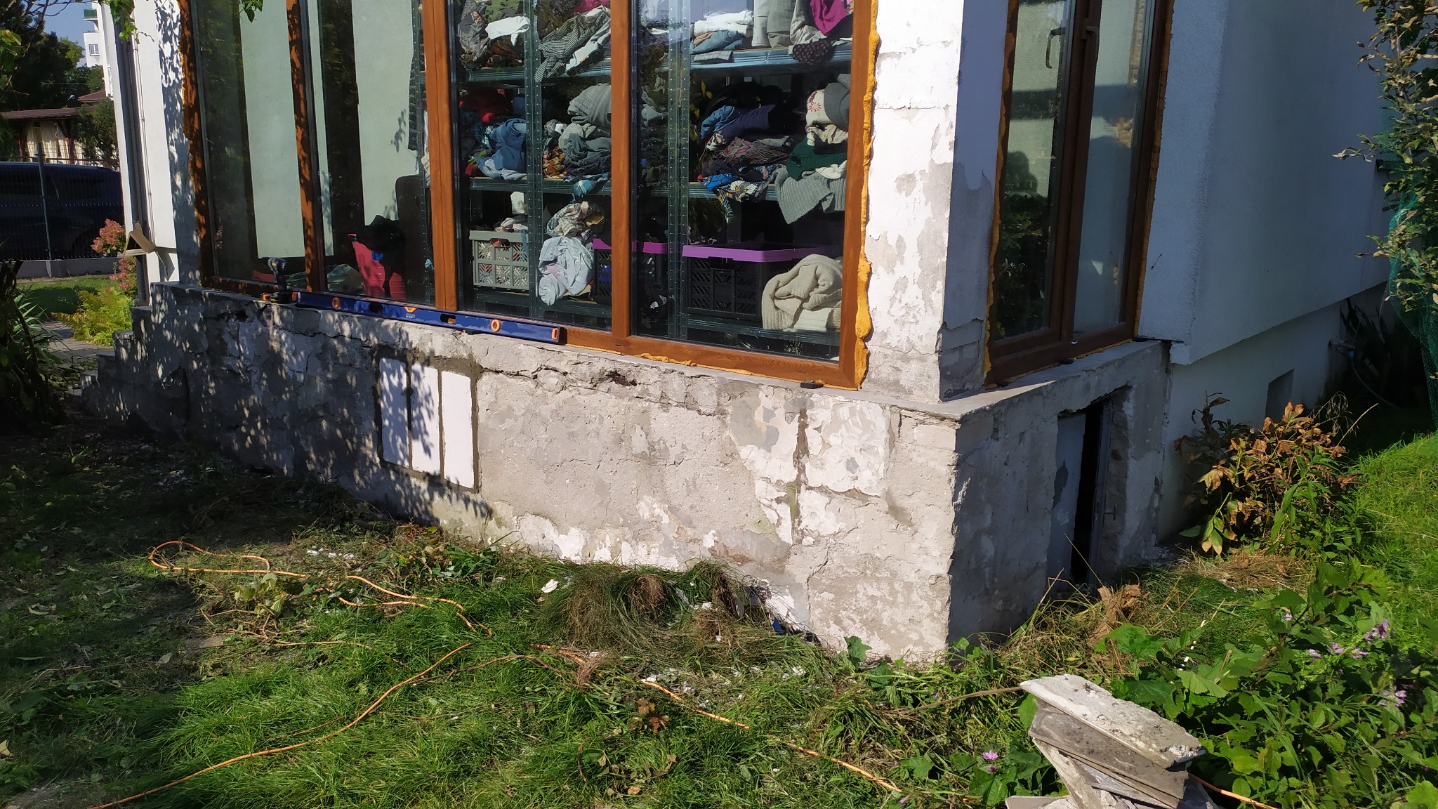 Fragment budynku z przeszkloną werandą, widoczne ślady prac remontowych na elewacji i nieporządek wokół budynku, poziomica przy oknie.