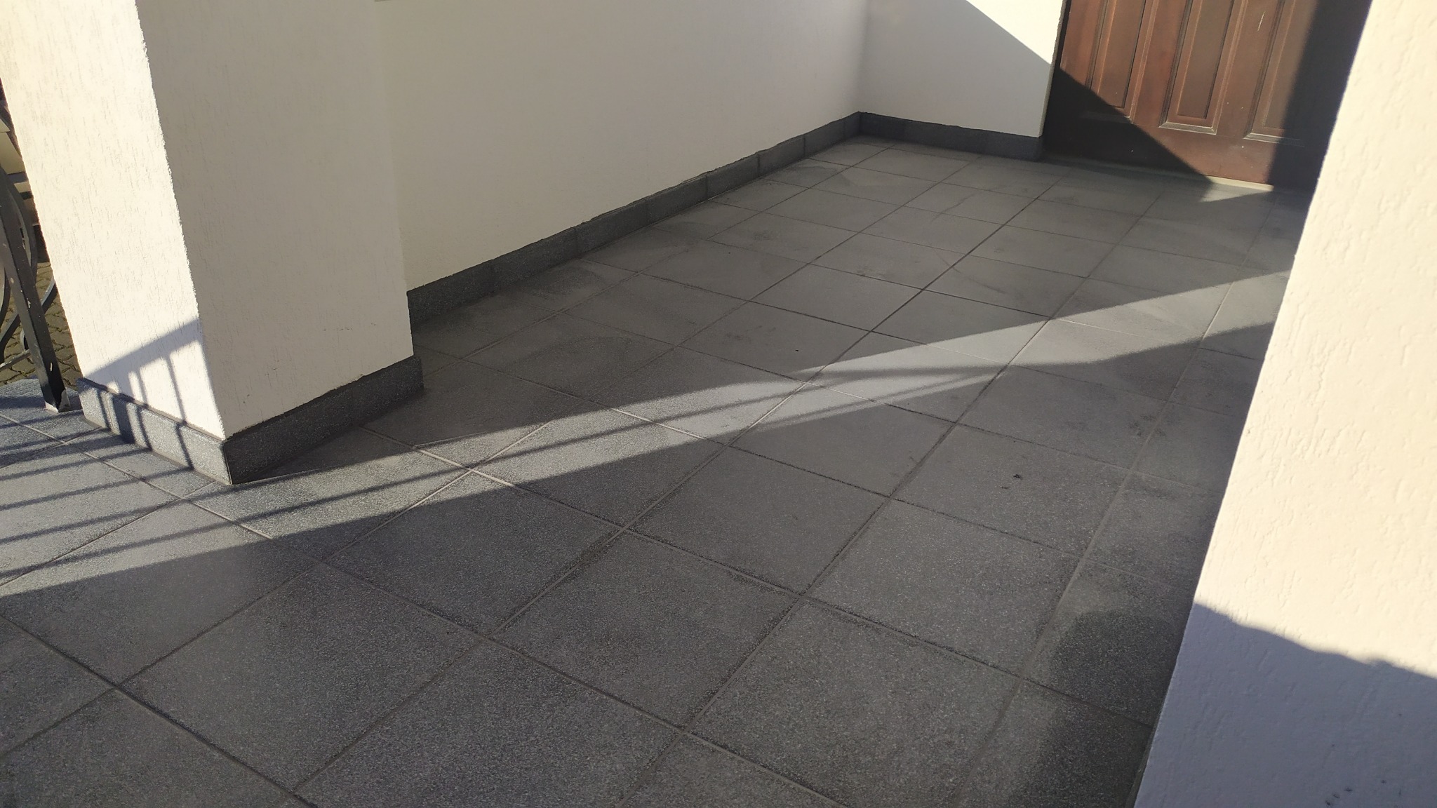 Wykończony balkon z szarymi płytkami podłogowymi, widoczne fragmenty balustrady i drzwi balkonowych w słoneczny dzień.