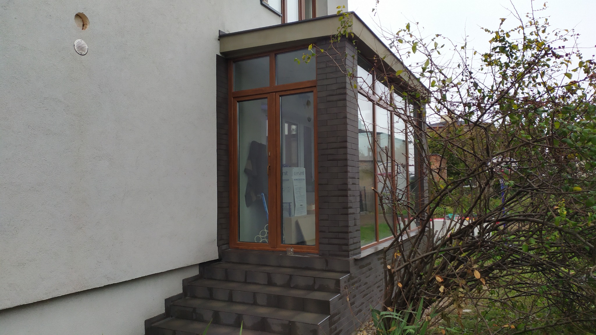 Zadaszone wejście do domu z brązowymi drzwiami i oknami, wykończone cegłą klinkierową, prowadzące po kilku schodach. Wewnątrz widoczne kartony i elementy wyposażenia.