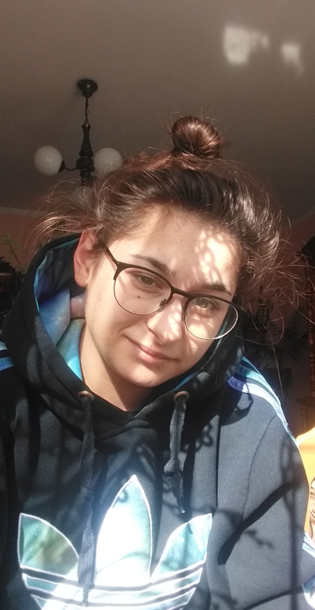 Portret uśmiechniętej młodej kobiety w okularach i czarnej bluzie z kapturem Adidas, z włosami upiętymi w kok na tle jasnego sufitu z żyrandolem.