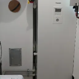 Wnętrze kotłowni z zamontowaną białą pompą ciepła Panasonic Aquarea, czerwonym i niebieskim zbiornikiem wyrównawczym, oraz elementami instalacji grzewczej, widoczne rury i osprzęt.