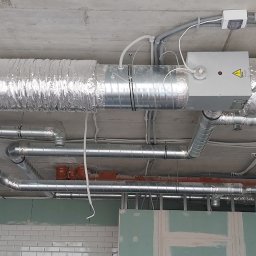 Instalklima - Srebrne rury wentylacyjne zamontowane pod betonowym sufitem w surowym stanie, widoczne przewody elektryczne i fragment ściany z białymi płytkami.