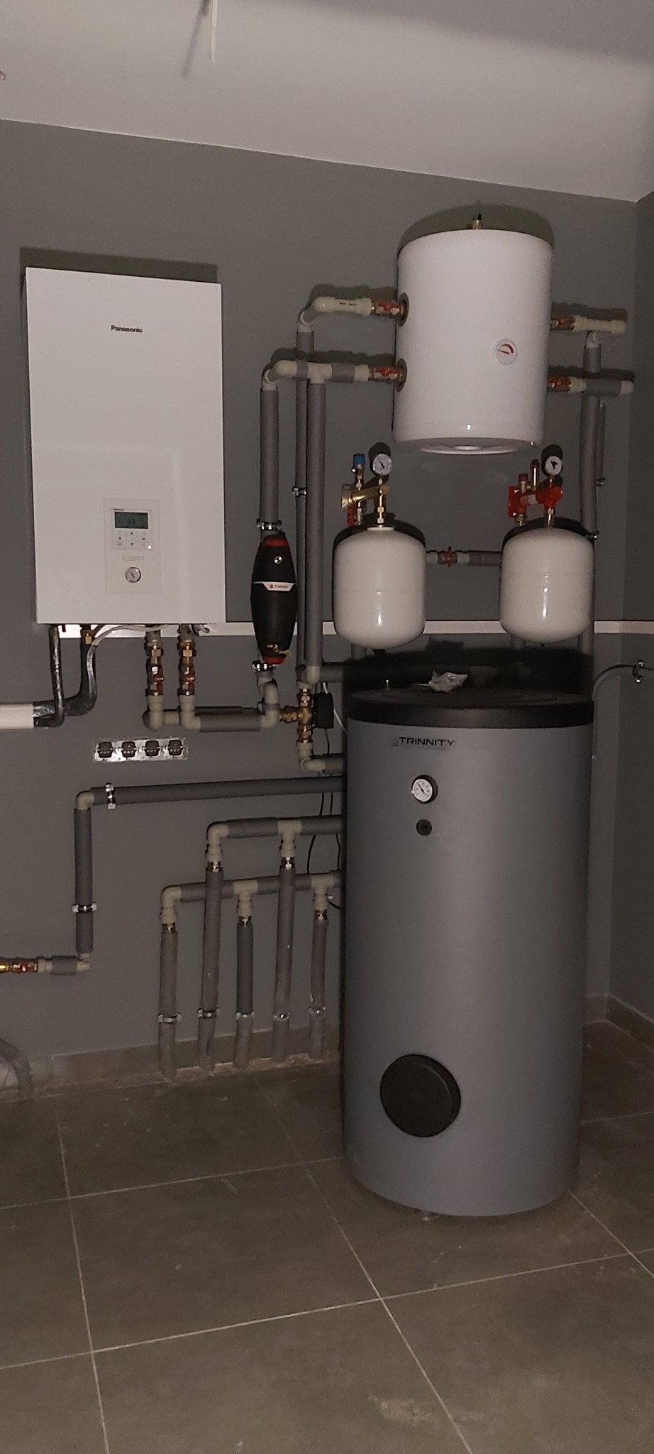 Zestaw instalacji grzewczej z piecem gazowym Panasonic i zasobnikiem Trinity, rury z izolacją, manometry, zamontowany na szarej ścianie w pomieszczeniu technicznym.