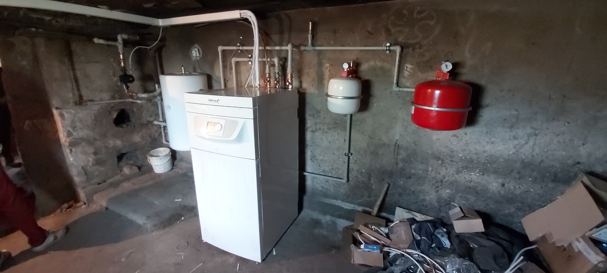 Nowy, biały piec gazowy marki Termet zainstalowany w piwnicy z surowymi, betonowymi ścianami. Widoczne rury, zbiornik wyrównawczy i bałagan po instalacji.