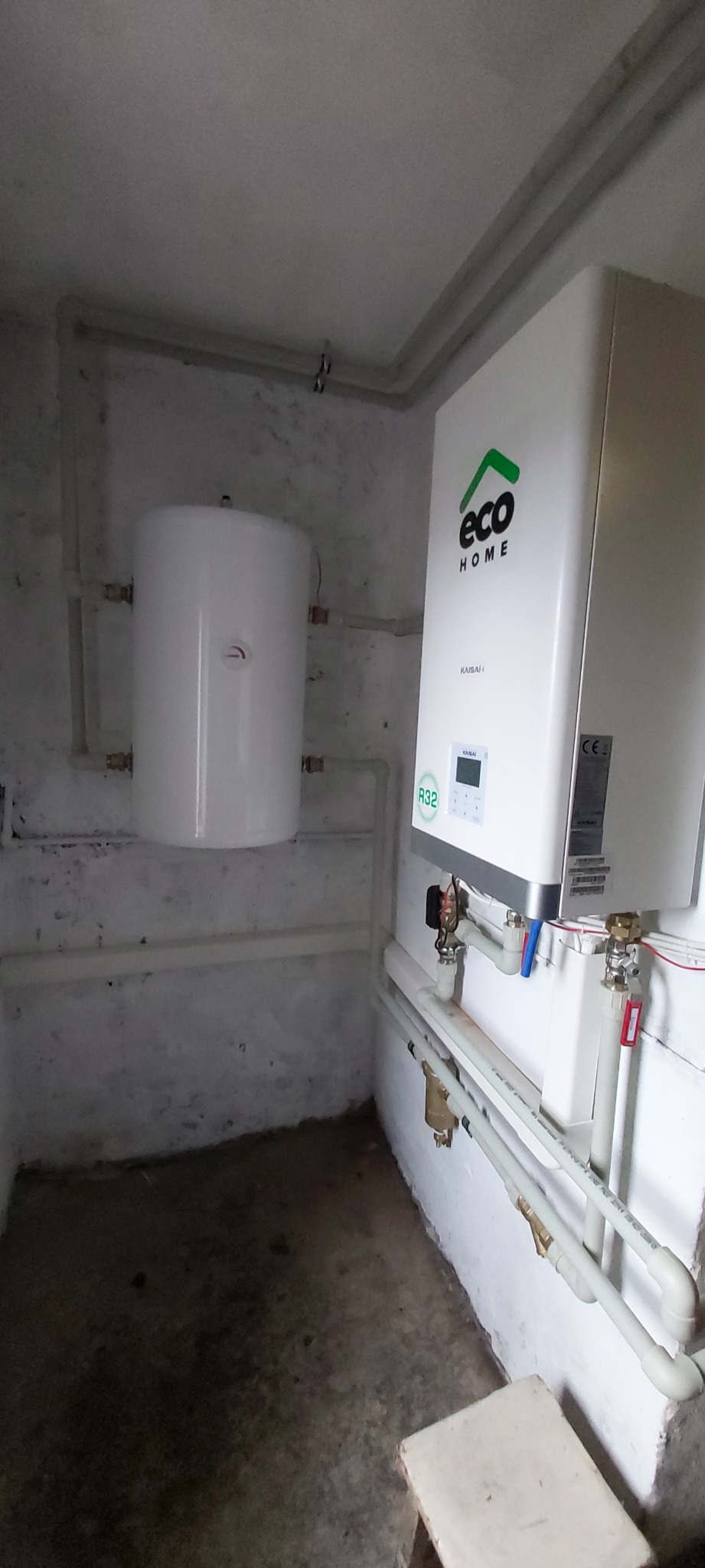 Biały piec gazowy marki Eco Home Kaisai z podłączonym zasobnikiem CWU, zamontowany na pomalowanej na biało ścianie w piwnicy, z widocznymi rurami instalacyjnymi.