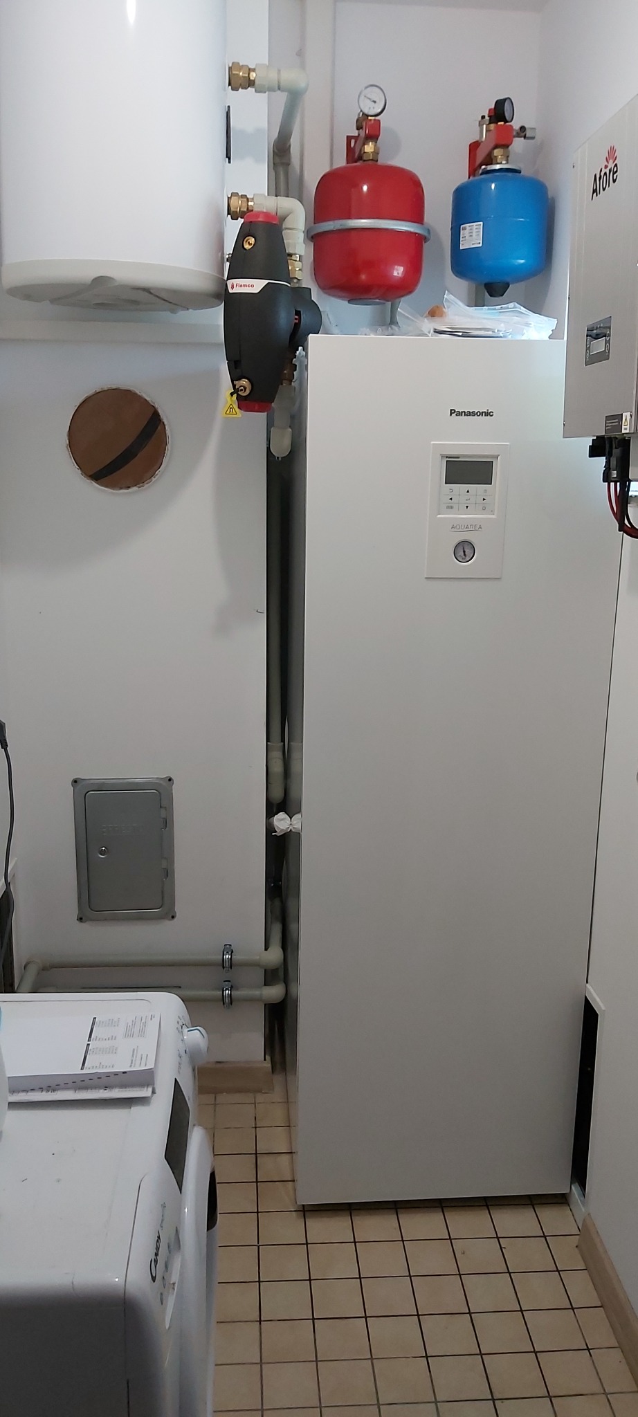 Wnętrze kotłowni z zamontowaną białą pompą ciepła Panasonic Aquarea, czerwonym i niebieskim zbiornikiem wyrównawczym, oraz elementami instalacji grzewczej, widoczne rury i osprzęt.