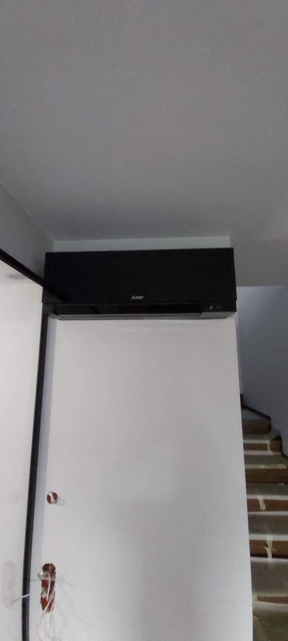 Czarny klimatyzator Mitsubishi zamontowany na białej ścianie w pomieszczeniu, widoczne niedokończone instalacje elektryczne i fragment schodów w tle.