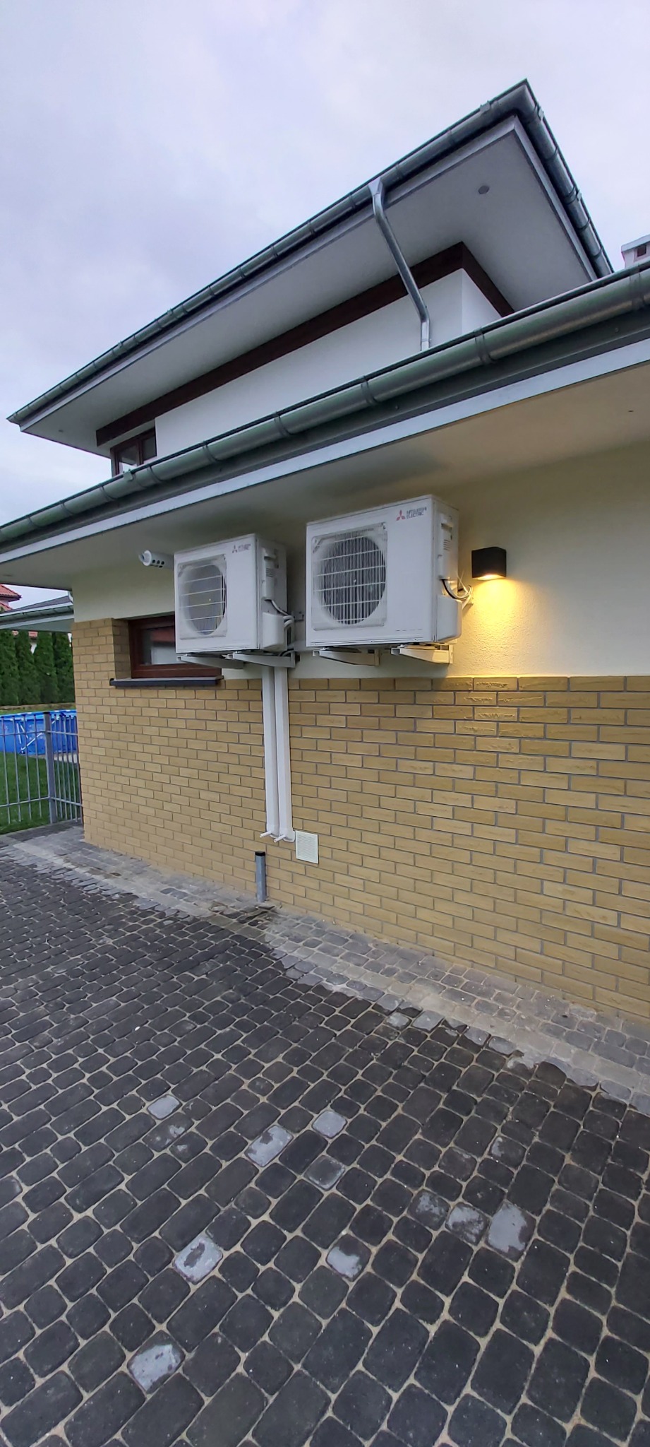 Dwa białe klimatyzatory Mitsubishi zamontowane na ścianie budynku z żółtej cegły, widoczne rury odprowadzające skropliny, oświetlenie zewnętrzne i kostka brukowa na podłożu.