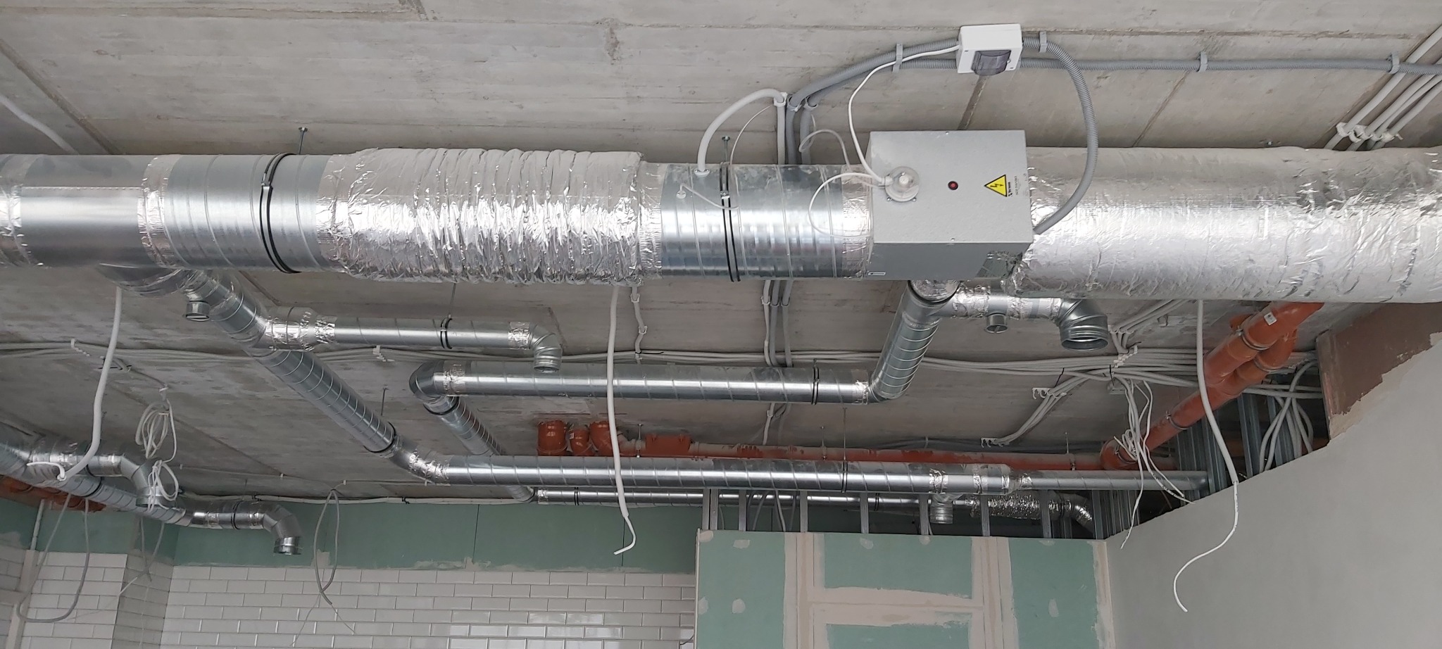 Srebrne rury wentylacyjne zamontowane pod betonowym sufitem w surowym stanie, widoczne przewody elektryczne i fragment ściany z białymi płytkami.