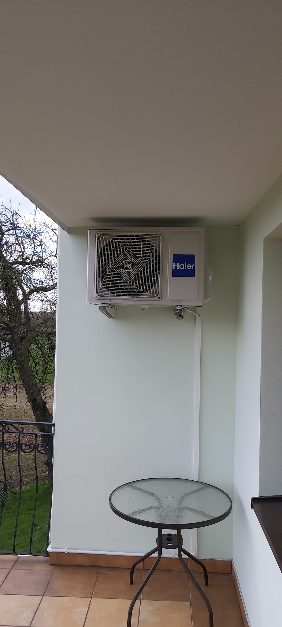 Biała jednostka klimatyzacyjna Haier zamontowana na ścianie balkonu, z widocznymi przewodami i stolikiem z blatem ze szkła w tle.