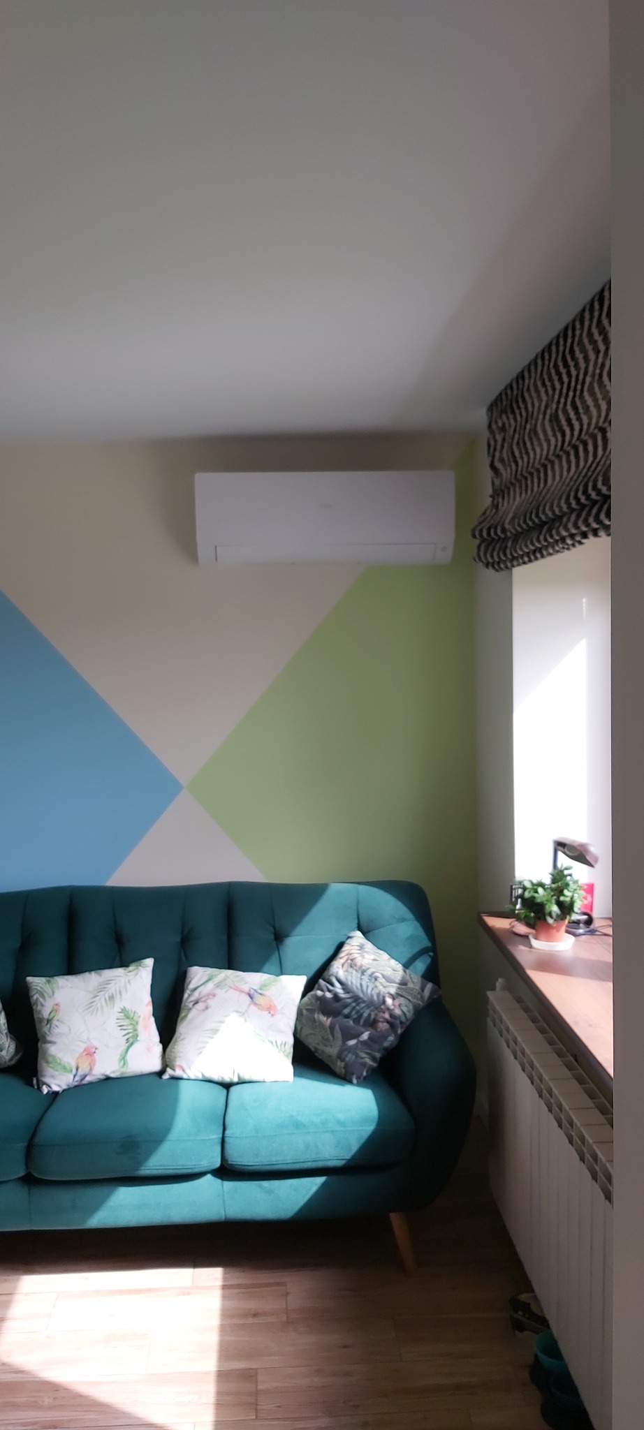 Wnętrze pokoju z turkusową sofą, pod oknem grzejnik, na ścianie klimatyzator i dekoracyjne malowanie w geometryczne wzory.