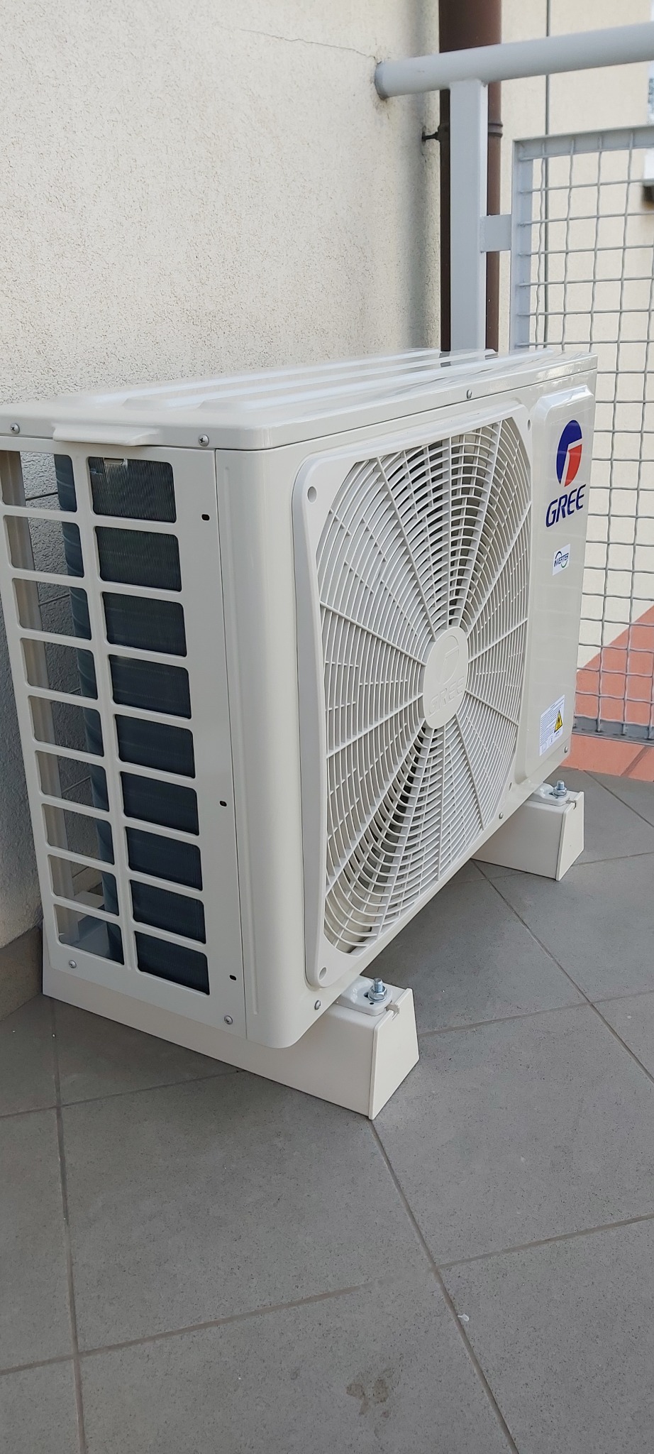 Zewnętrzna jednostka klimatyzatora marki Gree zamontowana na balkonie, widok z boku, z widocznym grillem wentylacyjnym i logo producenta.
