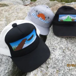 Trzy czapki z daszkiem leżące na szarym kamieniu: czarna z naszywką przedstawiającą górski krajobraz, szara z napisem 'Hey Vacay!' i palmami oraz czarno-biała z naszywką przedstawiającą pomost.