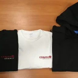 Trzy złożone ubrania na drewnianym stole: czarna bluza z kapturem, czarna bluza bez kaptura i biały t-shirt, wszystkie z wyhaftowanym czerwonym logo 'CZAJKA GRANIT'.
