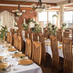 Uroczysta sala z długim stołem nakrytym białym obrusem, złotymi talerzami i dekoracjami kwiatowymi, z drewnianymi krzesłami o wysokich oparciach i ceglaną ścianą w tle.