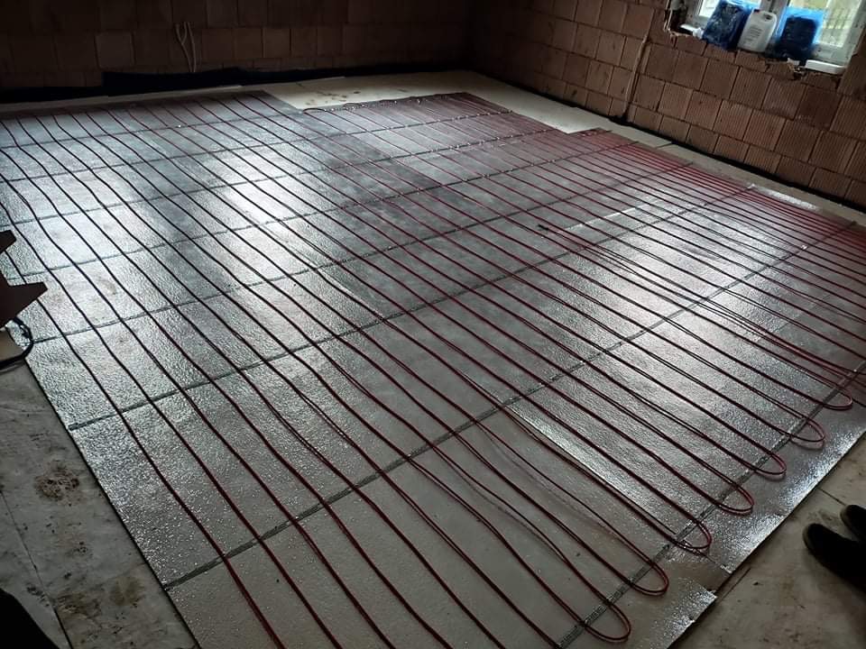 Instalacja ogrzewania podłogowego z czerwonymi przewodami na srebrnej folii izolacyjnej, widoczne fragmenty surowych ścian z cegły i okno z pojemnikami w tle.