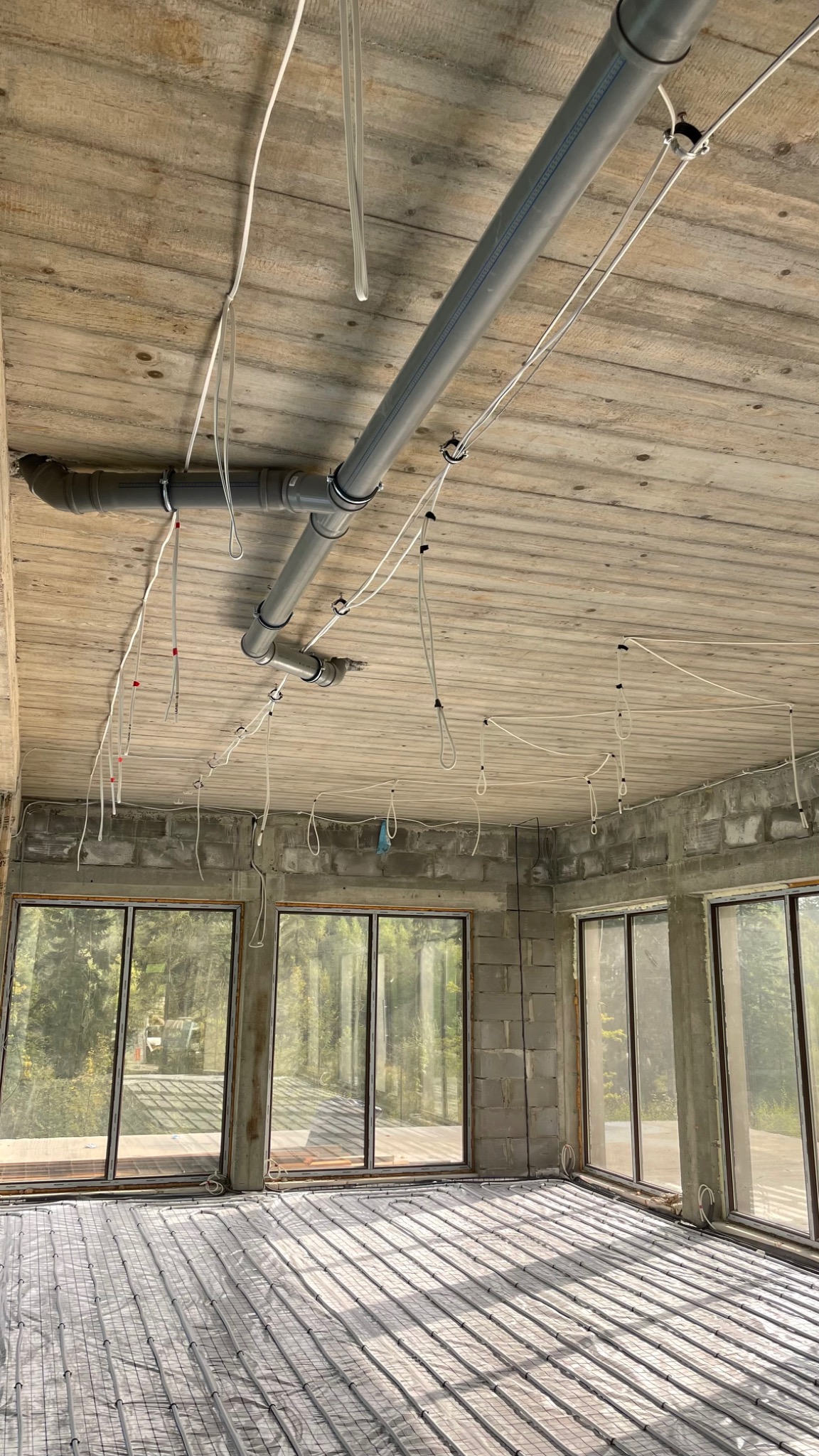 Surowy stan wnętrza z widocznymi przewodami elektrycznymi zwisającymi z sufitu i rurami kanalizacyjnymi, ogrzewanie podłogowe w trakcie montażu, duże okna z widokiem na zieleń.