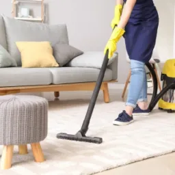 Osoba w ż&oacute;łtych rękawicach i niebieskim fartuchu odkurza jasny dywan w salonie, widoczny szary puf, sofa z poduszkami i ż&oacute;łty odkurzacz.