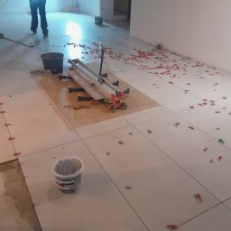 Pro-gres - Układanie jasnych płytek podłogowych z użyciem klipsów poziomujących i przecinarki do płytek. Fragment prac glazurniczych w pomieszczeniu.