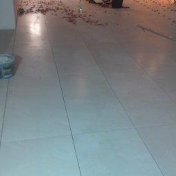 Pro-gres - Układanie jasnych, kwadratowych płytek podłogowych w dużej przestrzeni. Widoczne narzędzia glazurnicze, poziomica i pracownik w tle.