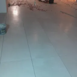 Układanie jasnych, kwadratowych płytek podłogowych w dużej przestrzeni. Widoczne narzędzia glazurnicze, poziomica i pracownik w tle.