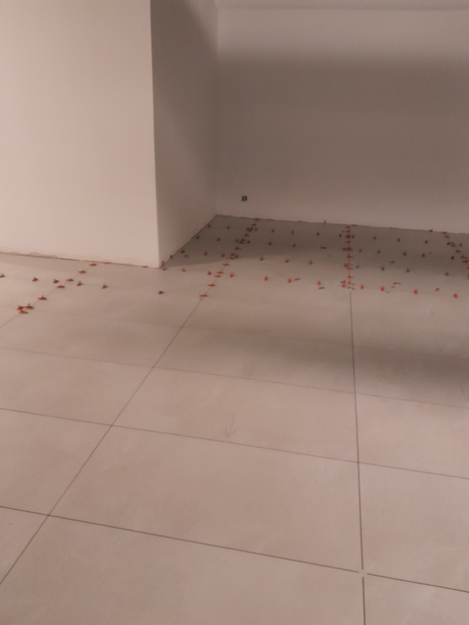 Ułożone, jasne płytki podłogowe w rogu pomieszczenia, z widocznymi pomarańczowymi krzyżykami dystansowymi. Widoczny fragment białej ściany.