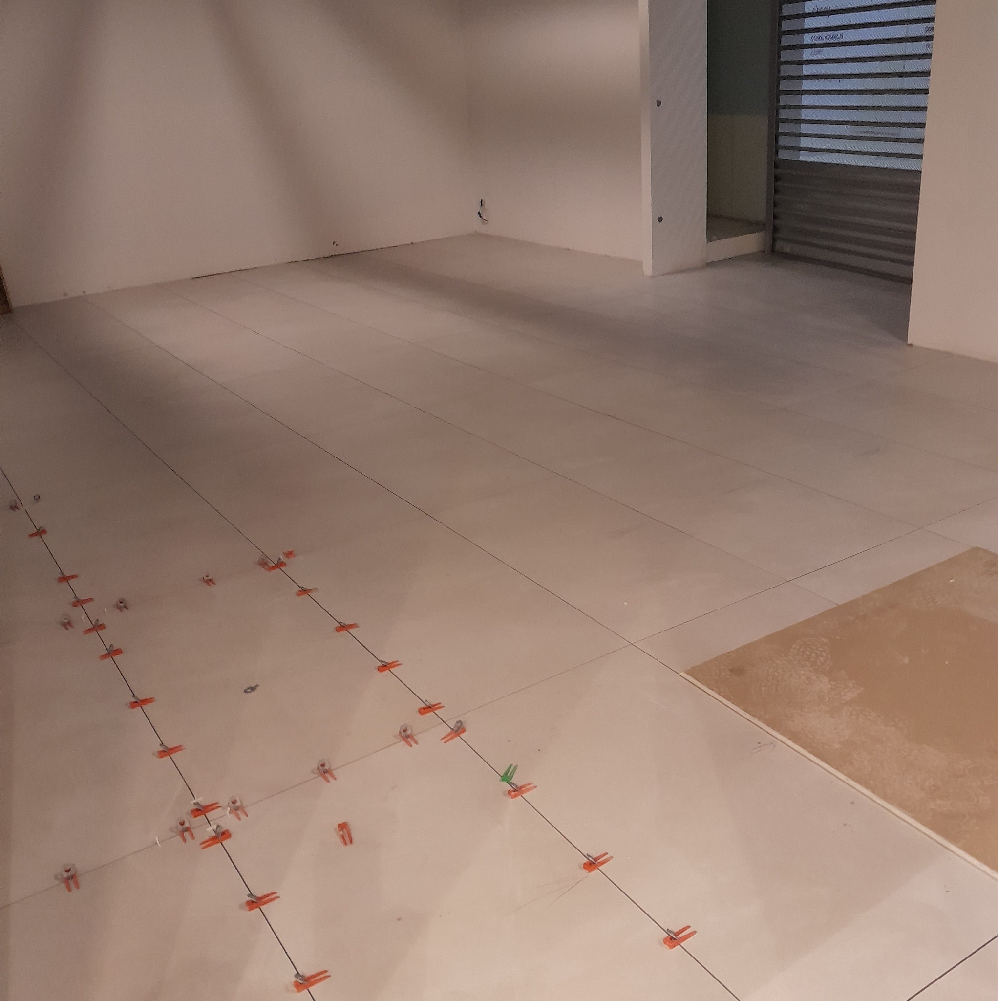 Układanie jasnych płytek podłogowych w pomieszczeniu, widoczne krzyżyki dystansowe i fragment niezabezpieczonej podłogi. Etap prac glazurniczych w jasnym wnętrzu.