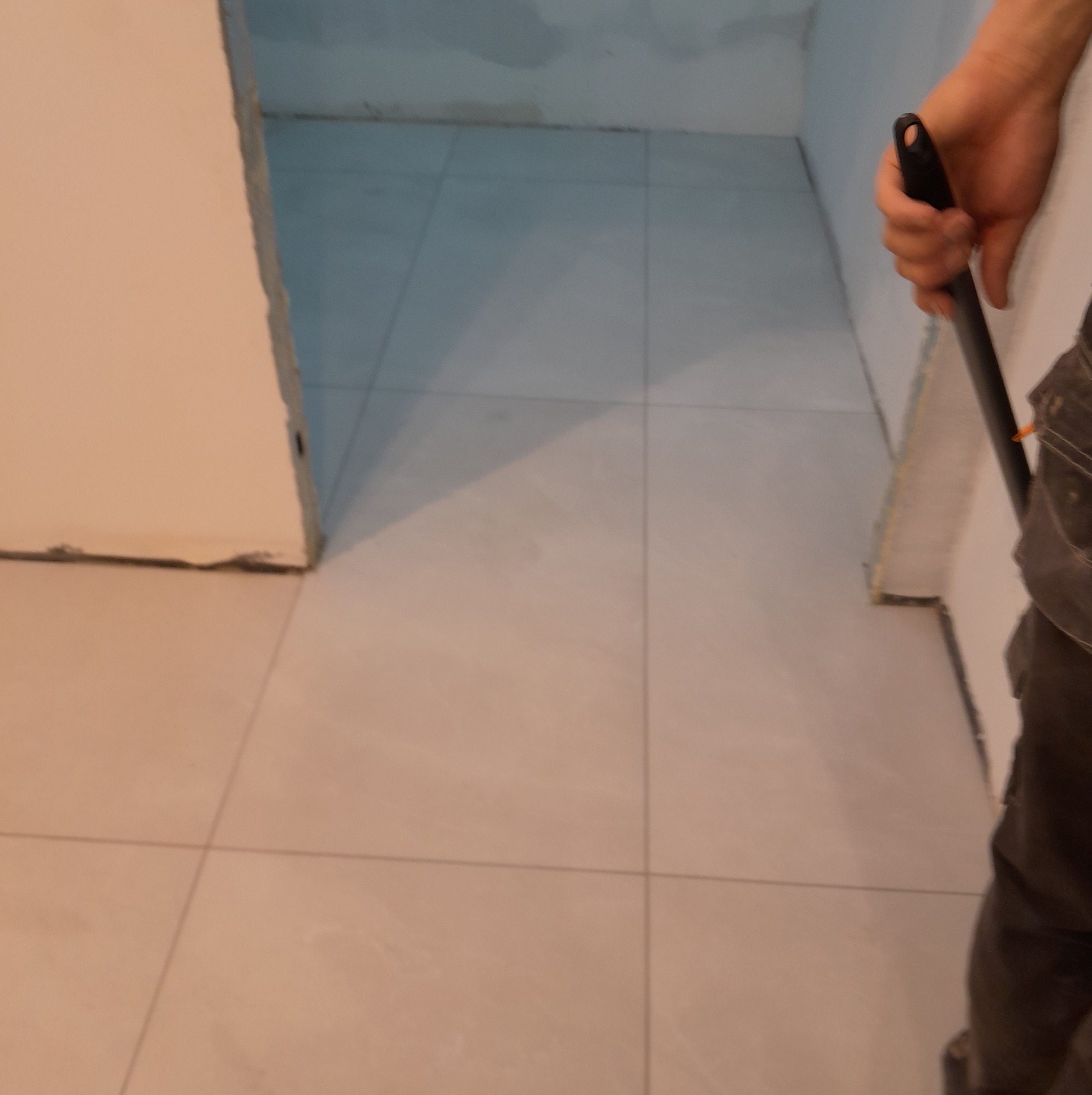 Jasne, nowo położone płytki podłogowe w pomieszczeniu. W tle widoczne ślady po szpachlowaniu na ścianach i fragment osoby trzymającej narzędzie.