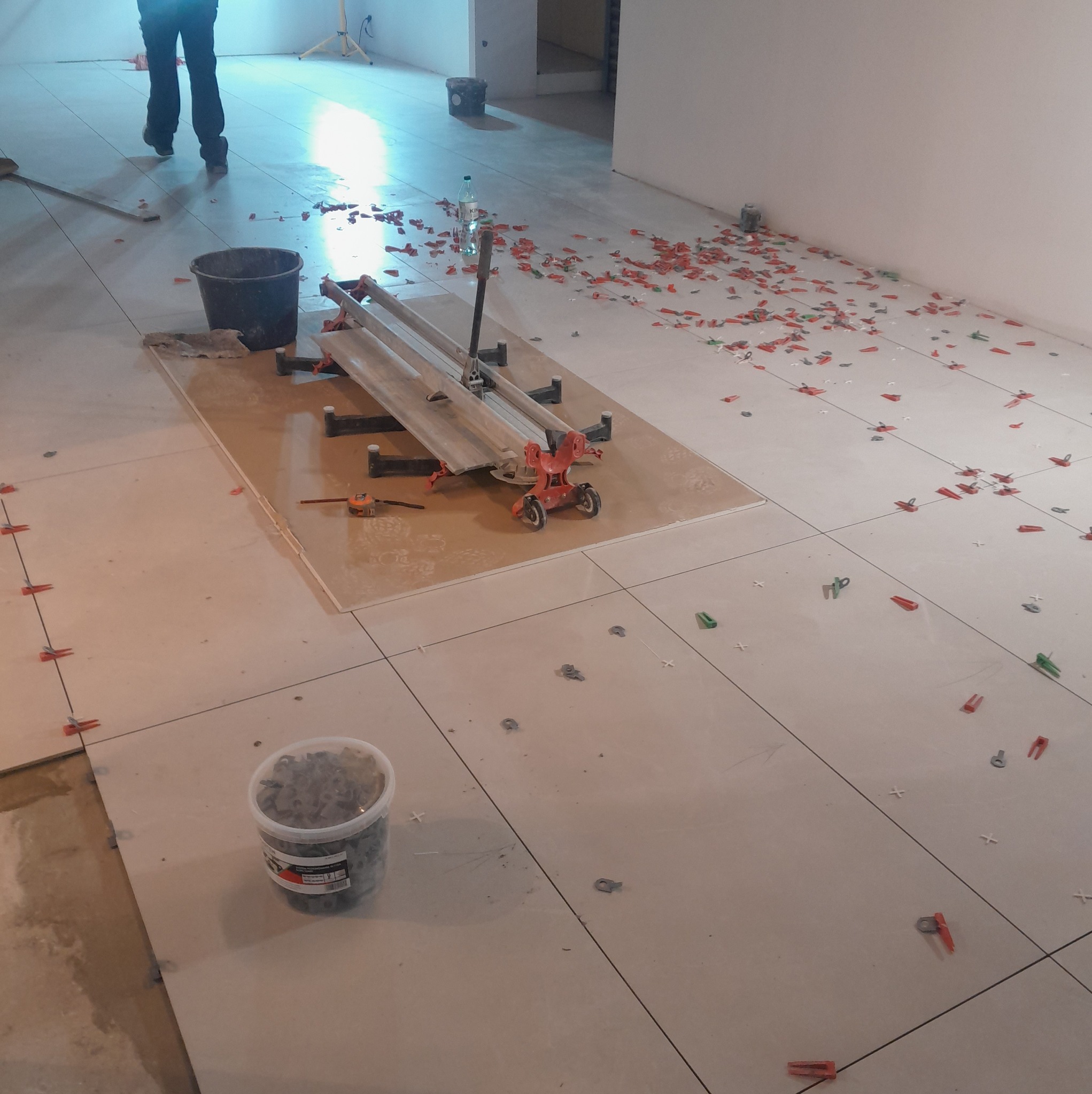 Układanie jasnych płytek podłogowych z użyciem klipsów poziomujących i przecinarki do płytek. Fragment prac glazurniczych w pomieszczeniu.