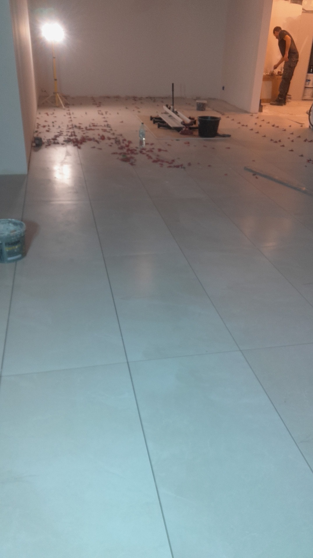 Układanie jasnych, kwadratowych płytek podłogowych w dużej przestrzeni. Widoczne narzędzia glazurnicze, poziomica i pracownik w tle.