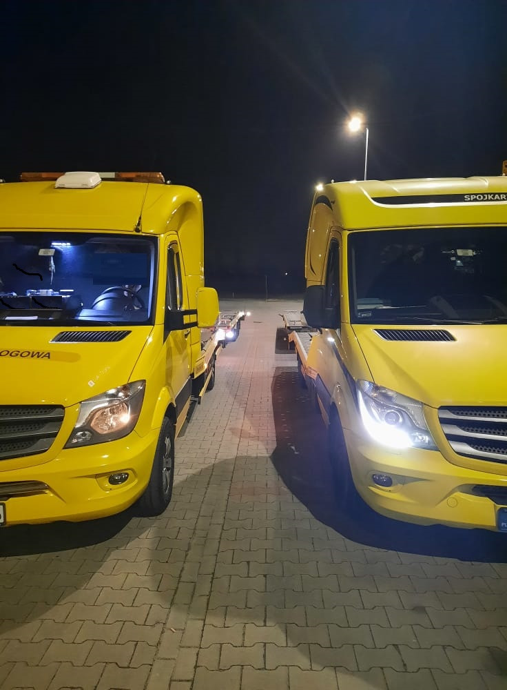 Dwa żółte samochody lawety Mercedes Sprinter stoją obok siebie na parkingu w nocy, oświetlone światłem latarni, z widocznymi napisami 'OGOWA' i 'SPOJKAR' na nadwoziu.