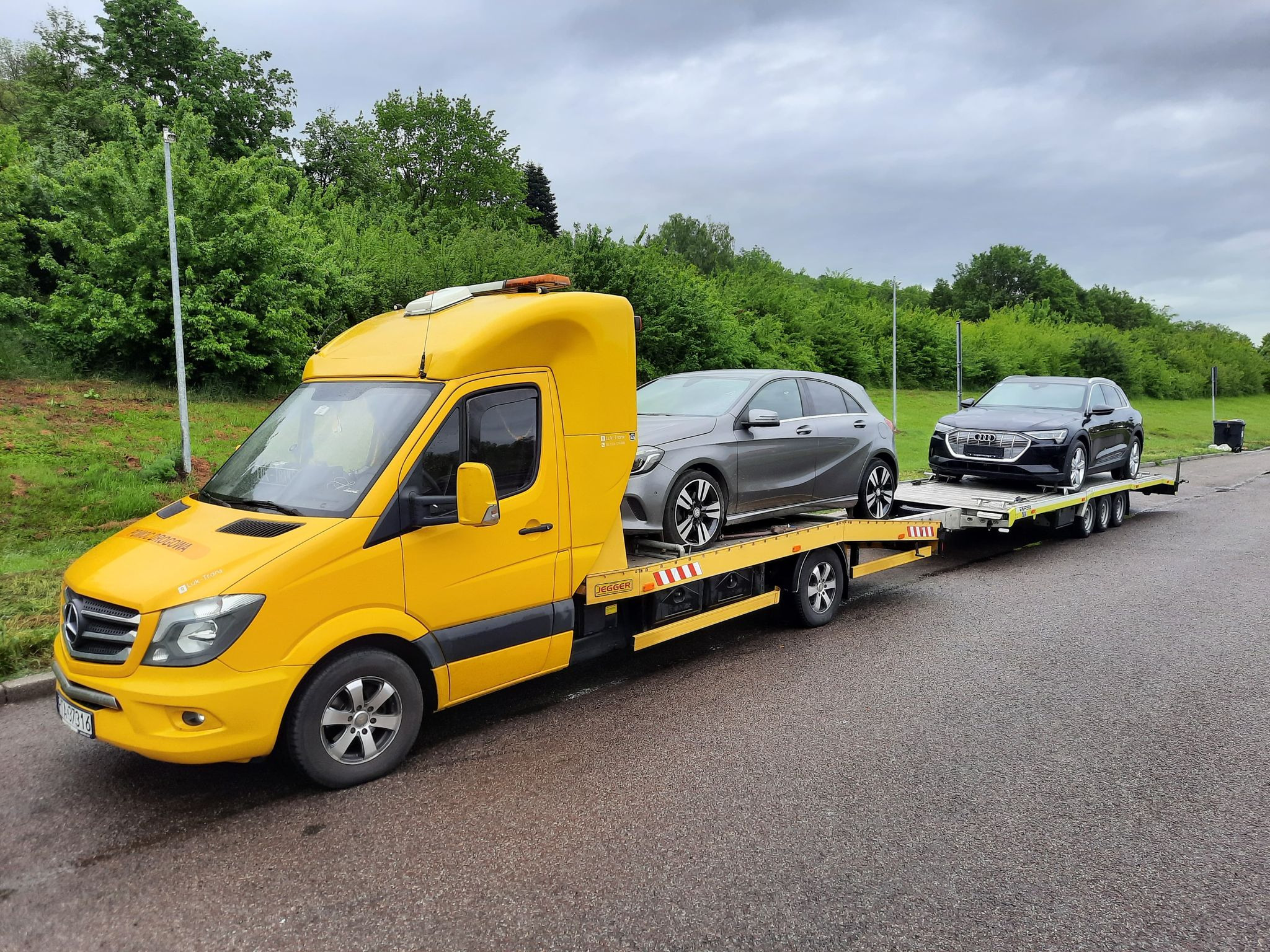 Żółty pojazd pomocy drogowej Mercedes-Benz z dwoma samochodami osobowymi na platformie, szary Mercedes i czarne Audi, na tle zieleni i pochmurnego nieba.