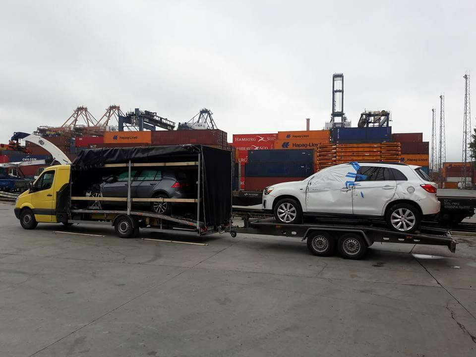 Żółty samochód dostawczy z przyczepą przewozi dwa samochody osobowe na tle portowych dźwigów i kontenerów, jeden z aut zabezpieczony folią.