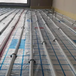 Rozłożone rury ogrzewania podłogowego na folii z nadrukiem SKALAR, przymocowane czarnymi klipsami, widoczne czerwone paski i żółta izolacja przy ścianie