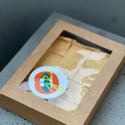 Kartonowe pudełko z okienkiem wypełnione kawałkami ciasta z białym lukrem, ozdobione naklejką z abstrakcyjnym logo w kolorach pomarańczowym, zielonym i żółtym.