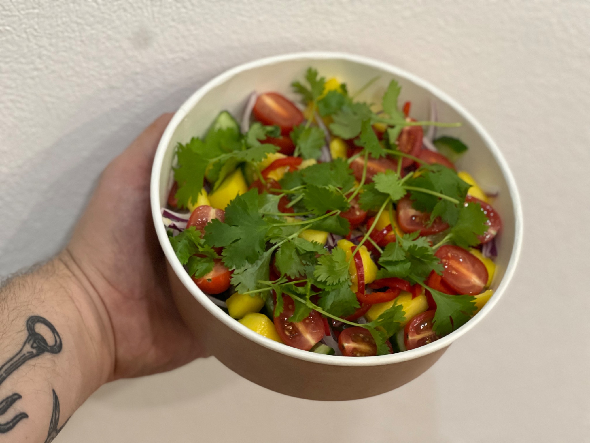 Sałatka z mango, pomidorków cherry, czerwonej cebuli, ogórka i kolendry w papierowej misce trzymana w dłoni z tatuażem kotwicy.