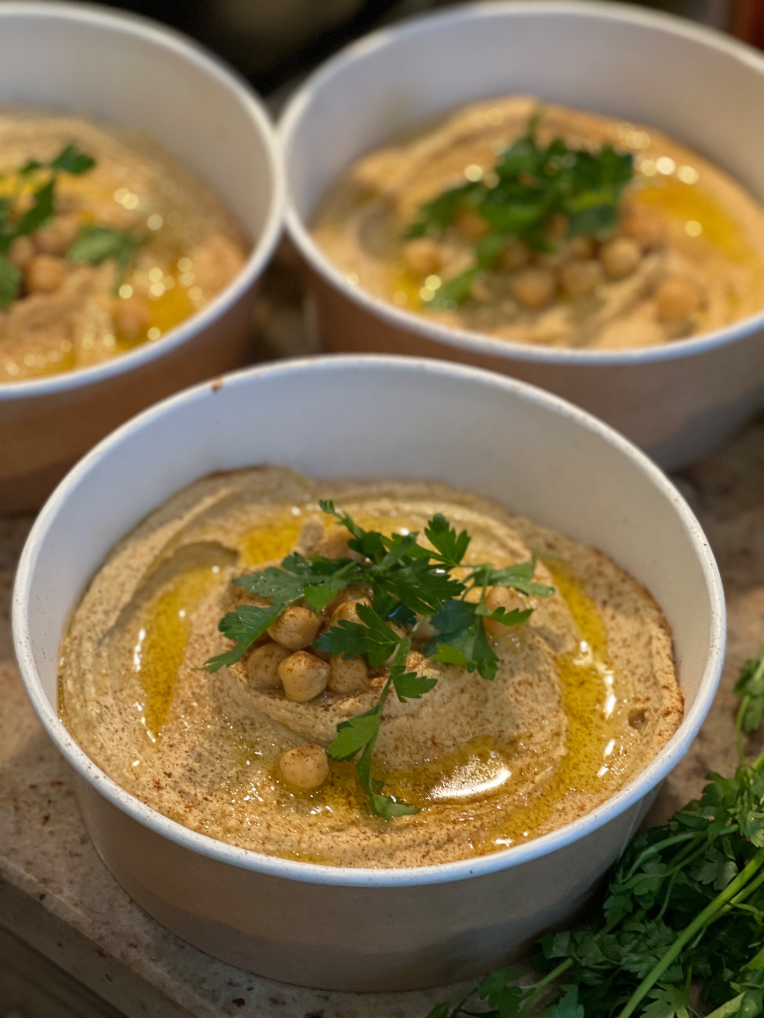 Trzy porcje kremowego hummusu w papierowych miseczkach, udekorowane świeżą natką pietruszki i ciecierzycą, polane oliwą, na blacie o teksturze kamienia.