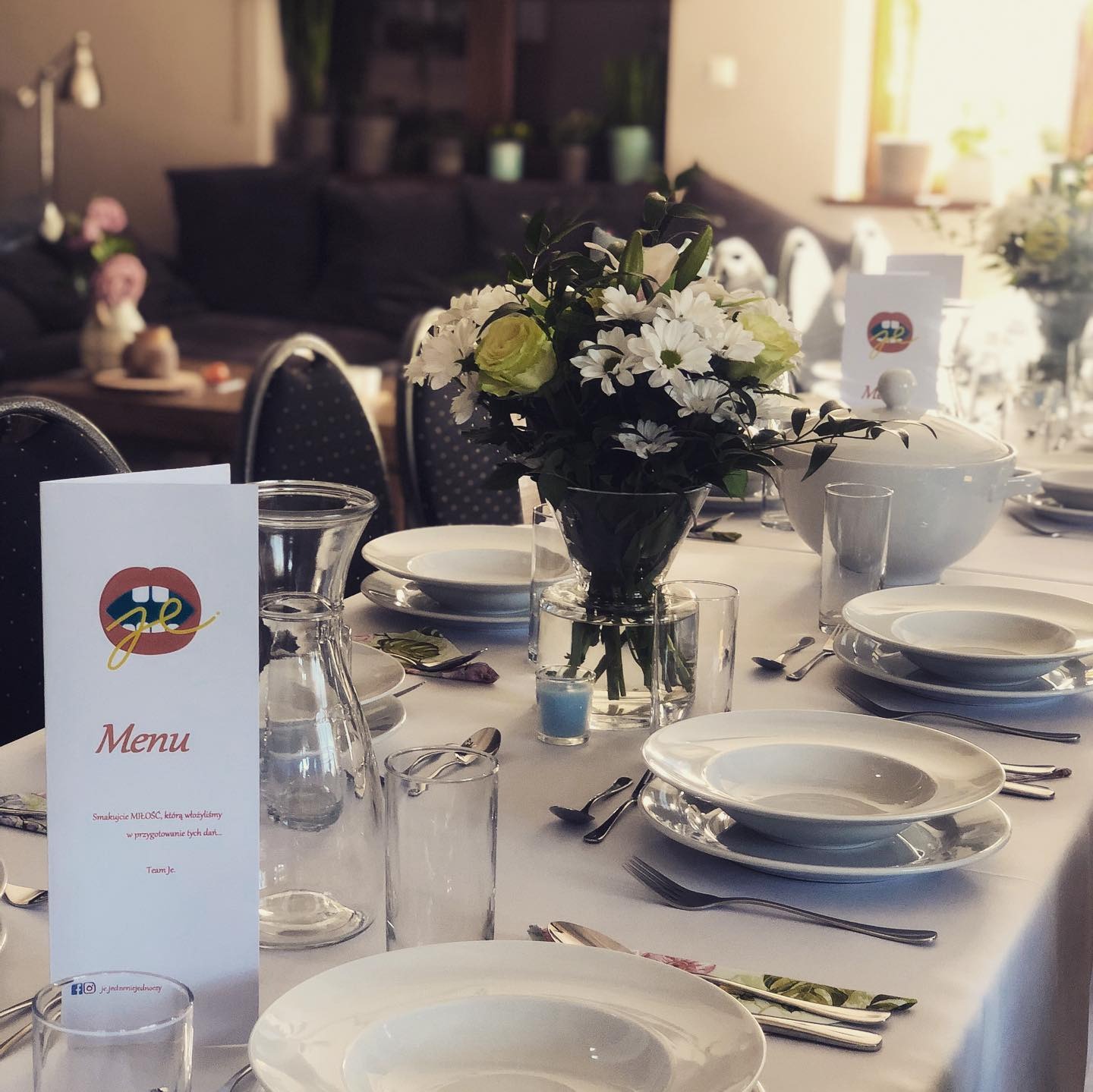 Elegancko nakryty stół z białym obrusem, zastawą, kwiatami w wazonie i menu z logo firmy cateringowej, przygotowany na uroczystość.
