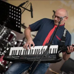 Muzyk grający na keytarze Roland AX-Synth podczas koncertu na scenie, w tle widoczne instrumenty perkusyjne i statyw z nutami.