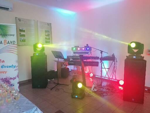 Scena oświetlona kolorowymi światłami, z klawiaturą, mikrofonem i nagłośnieniem, przygotowana do występu na imprezie, widoczny baner reklamowy z nazwą zespołu.
