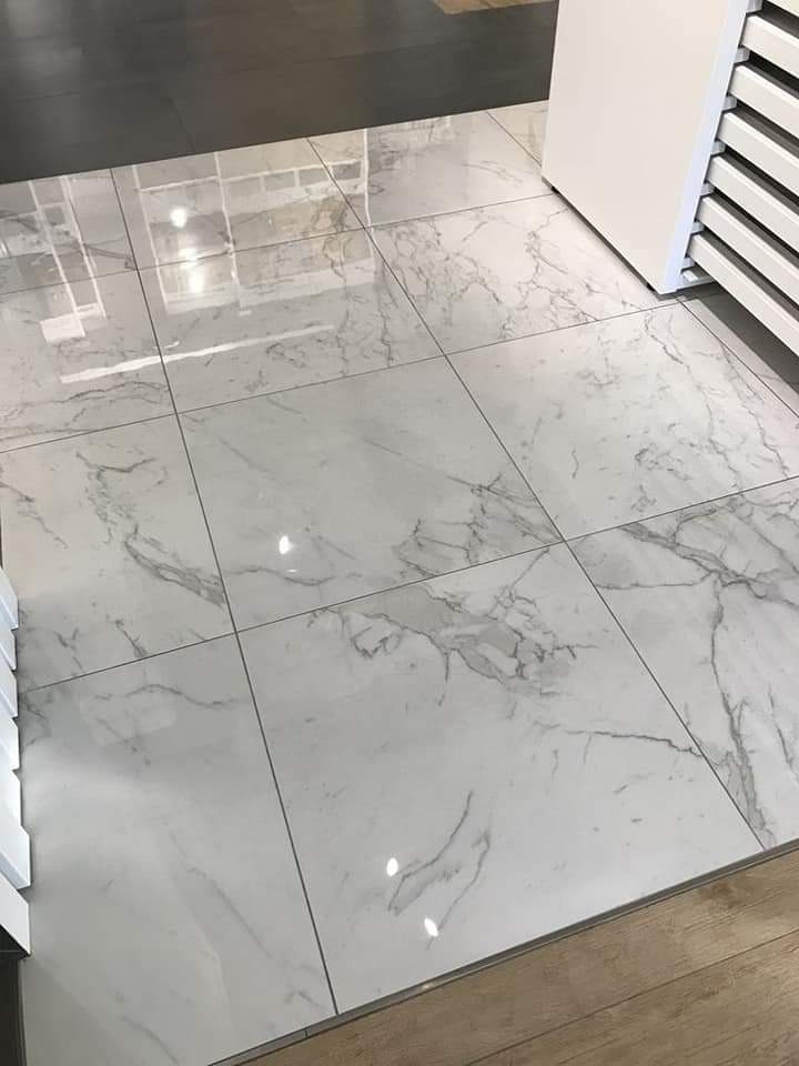 Białe, kwadratowe płytki podłogowe z szarym wzorem marmuru, ułożone z wąską fugą, widoczne odbicia światła, sąsiadują z drewnianą podłogą i białym regałem z próbkami.