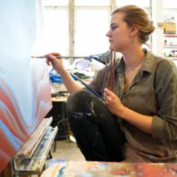 Artystka maluje abstrakcyjny obraz na płótnie w pracowni, trzymając pędzle w dłoni, widoczne okno w tle.