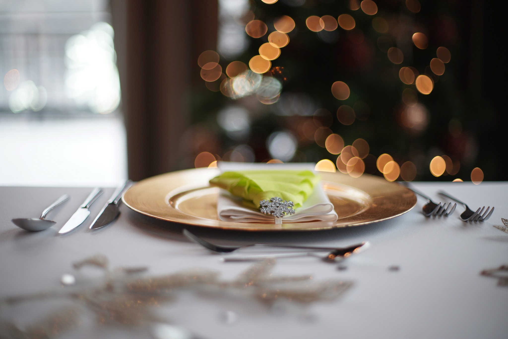 Elegancko nakryty stół z złotym talerzem, serwetką z ozdobnym pierścieniem i sztućcami; w tle rozmyte świąteczne lampki choinkowe.