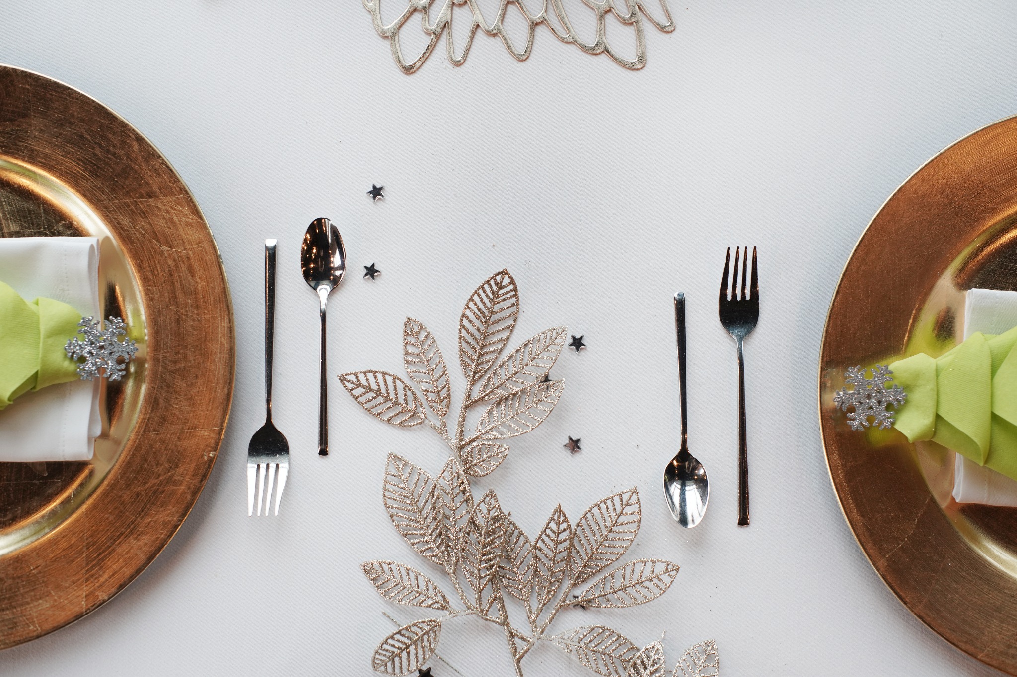 Uroczysta aranżacja stołu z dwoma złotymi talerzami, zielonymi serwetkami ozdobionymi srebrnymi płatkami śniegu, srebrnymi sztućcami i dekoracyjną gałązką z liśćmi w kolorze złota na białym tle.